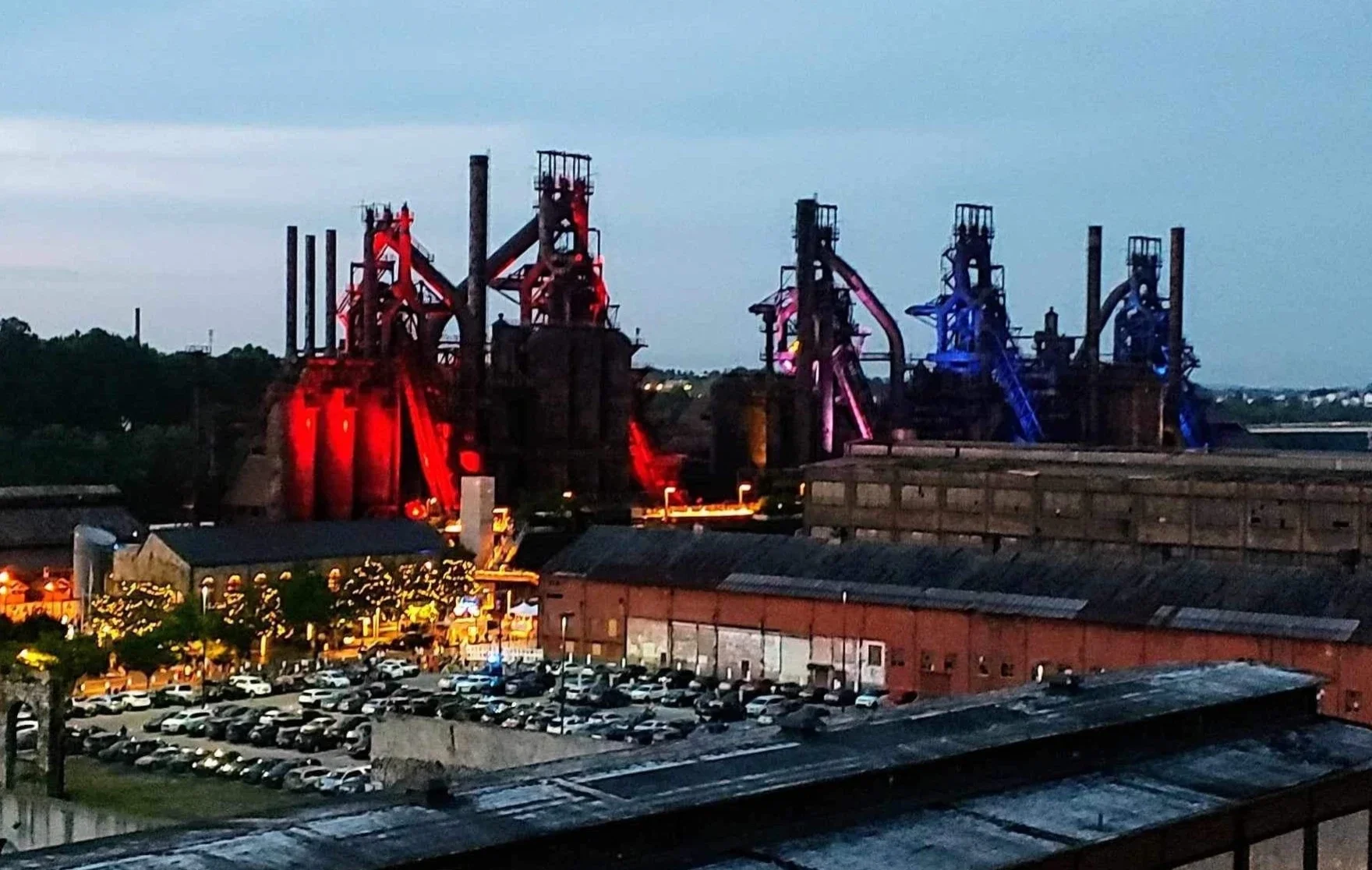 Industrial site with multiple large structures illuminated in red and blue lights, adjacent to a parking lot with many cars, at dusk.