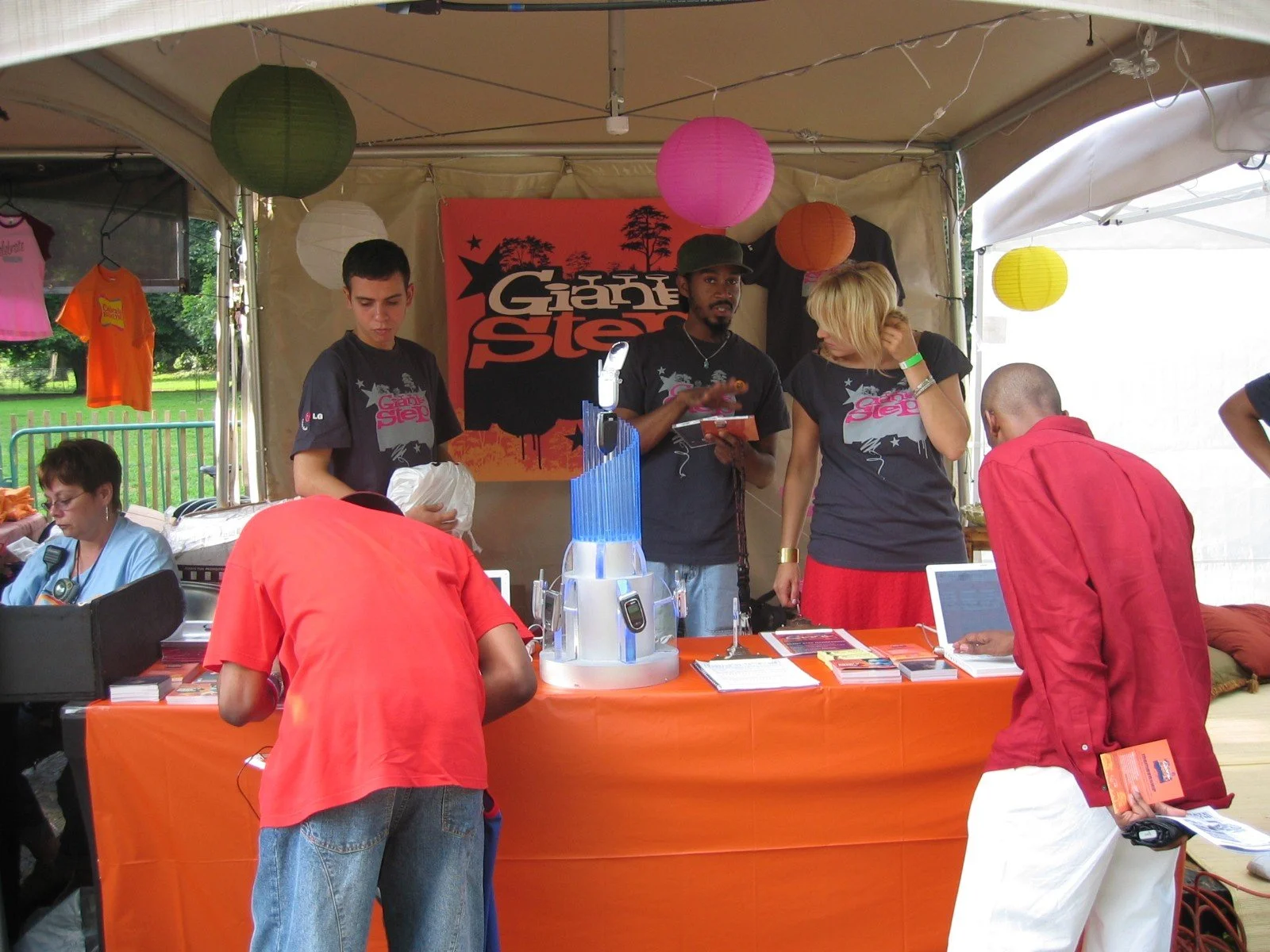 A group of people at a booth with an orange tablecloth, displaying flyers and pamphlets, under a canopy decorated with colorful paper lanterns. Three individuals are standing behind the booth, talking to visitors, with one person wearing a black t-sh