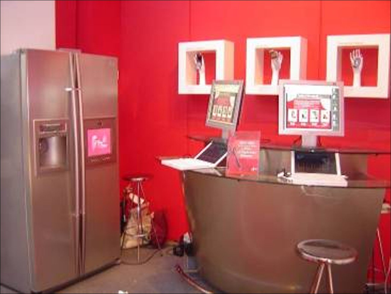 A red-themed customer service area with a stainless steel refrigerator, a curved counter, a computer monitor, and wall-mounted decorative frames with hand sculptures.