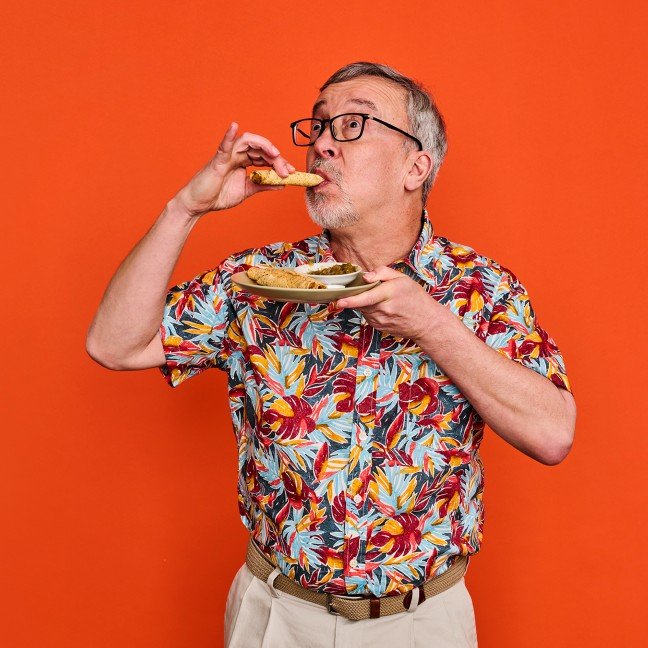 A middle-aged man with gray hair, glasses, and a goatee, wearing a colorful tropical shirt, holding a plate of food in one hand and eating a piece of bread or sandwich with the other, against an orange background.