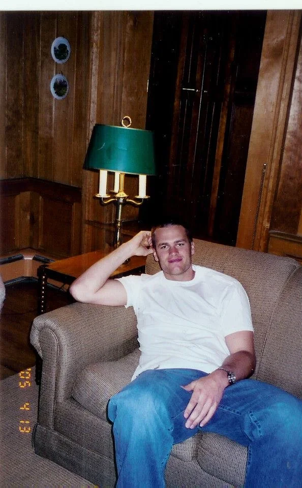 A young man relaxing on a beige sofa in a wood-paneled room, wearing a white t-shirt and blue jeans, with a green-shaded lamp and decorative plates on the wall behind him.