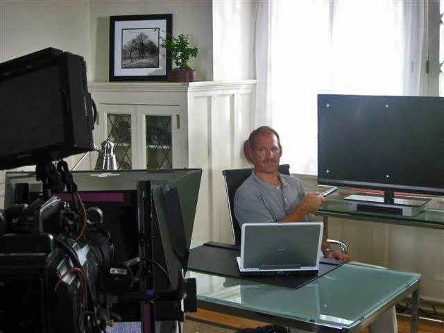 A man sitting at a glass desk with a laptop, holding a remote, in an office with a large monitor, a cabinet with glass doors, and a framed black-and-white picture on the wall.