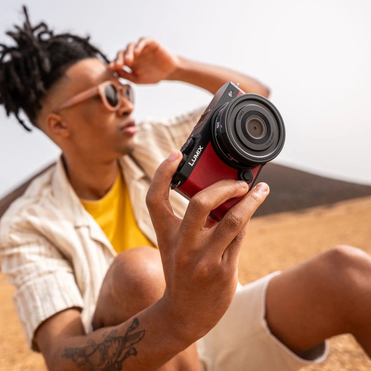 Person sitting on the floor holding a red Panasonic Lumix camera while looking at it, wearing sunglasses, a yellow shirt, and beige jacket.