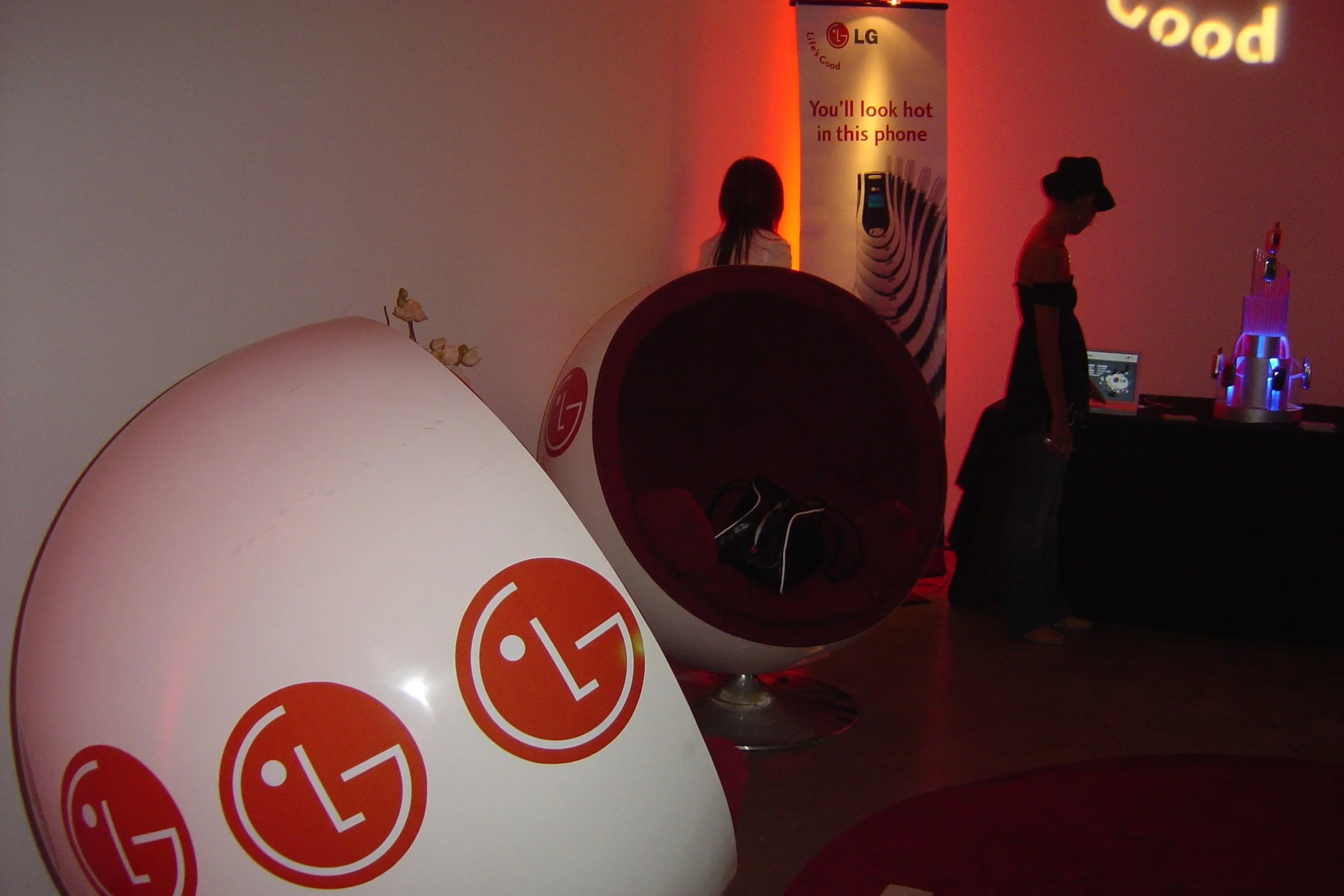 An LG promotional display area with a large LG logo illuminated on a sphere and other LG branding elements. There are modern, pod-like lounge chairs with LG logos, and a poster with the LG logo and slogan, "You'll look hot in this phone." In the back