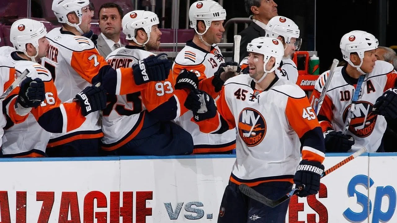 Ice hockey players from the New York Islanders team on the bench during a game, wearing white jerseys with orange and navy accents, with one player standing in front holding a hockey stick and smiling.