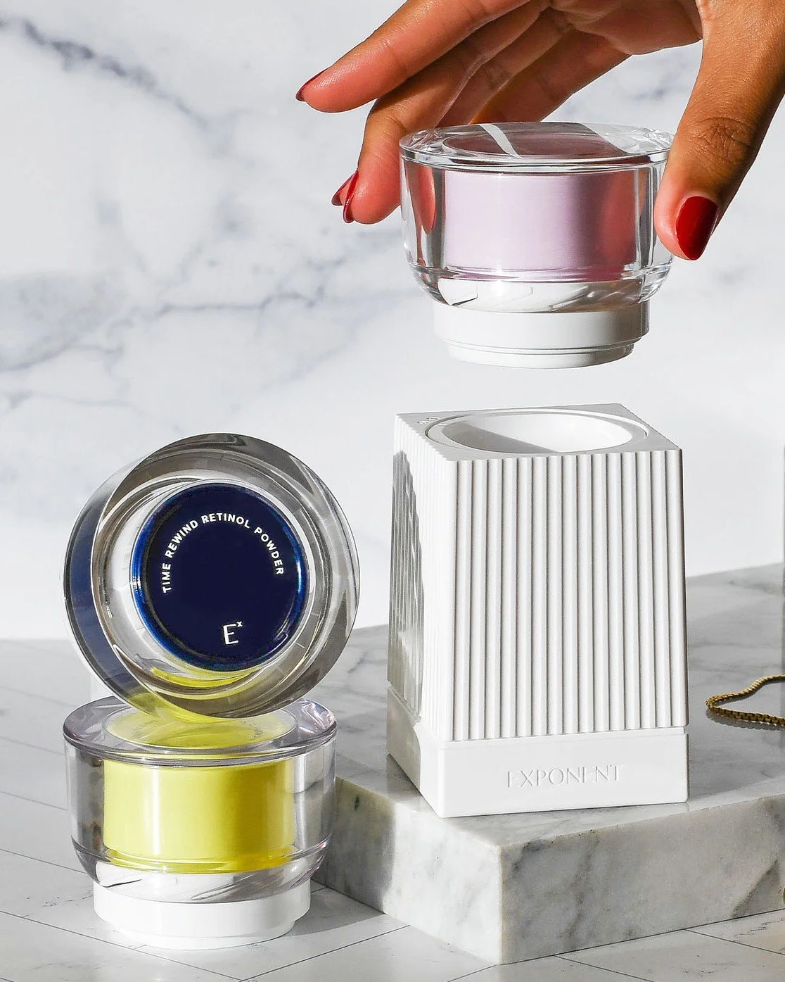A hand with red painted nails holding a clear jar above a stack of cosmetic containers on a white marble surface. The containers include a yellow cream and a silver-colored container with a dark blue label reading "Time Rewind Retinol Powder."