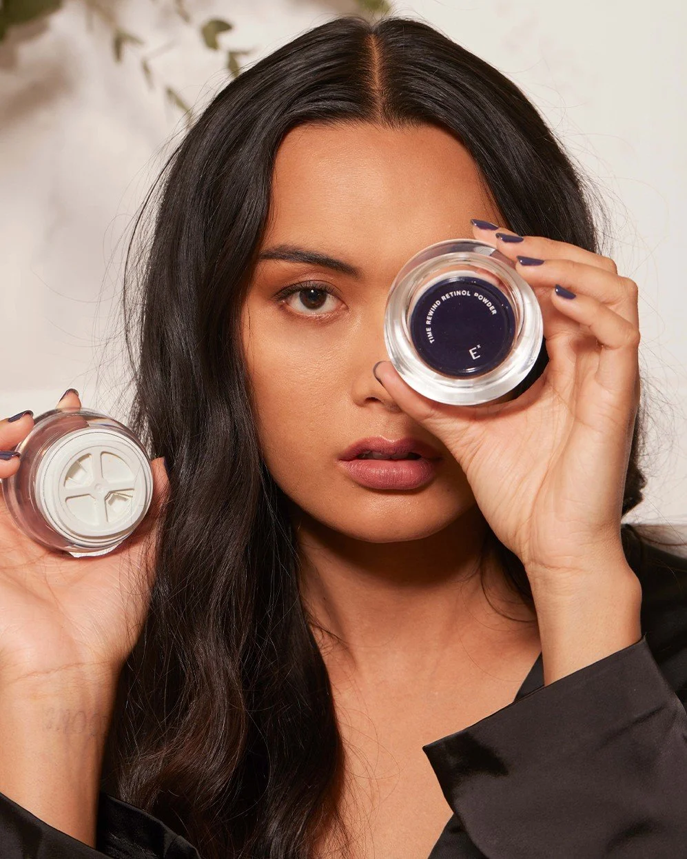 A woman with long dark hair holding a small jar of retinol powder up to her right eye, with her left eye looking at the camera. She is wearing a black satin top.