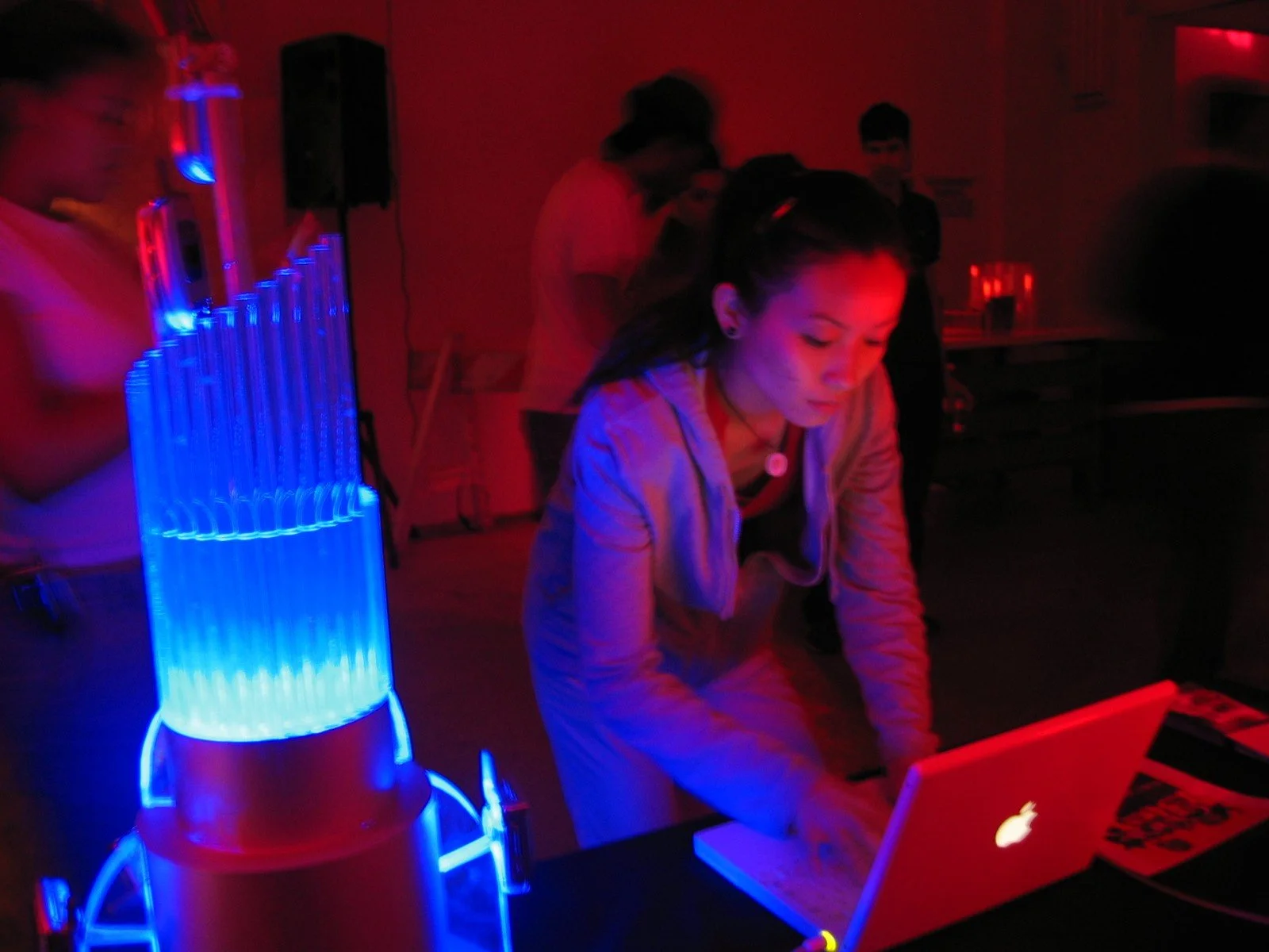A woman wearing a beige jacket using a white MacBook laptop at a table in a dimly lit room with red lights, with a blue and red water rocket in the foreground and a group of people in the background.
