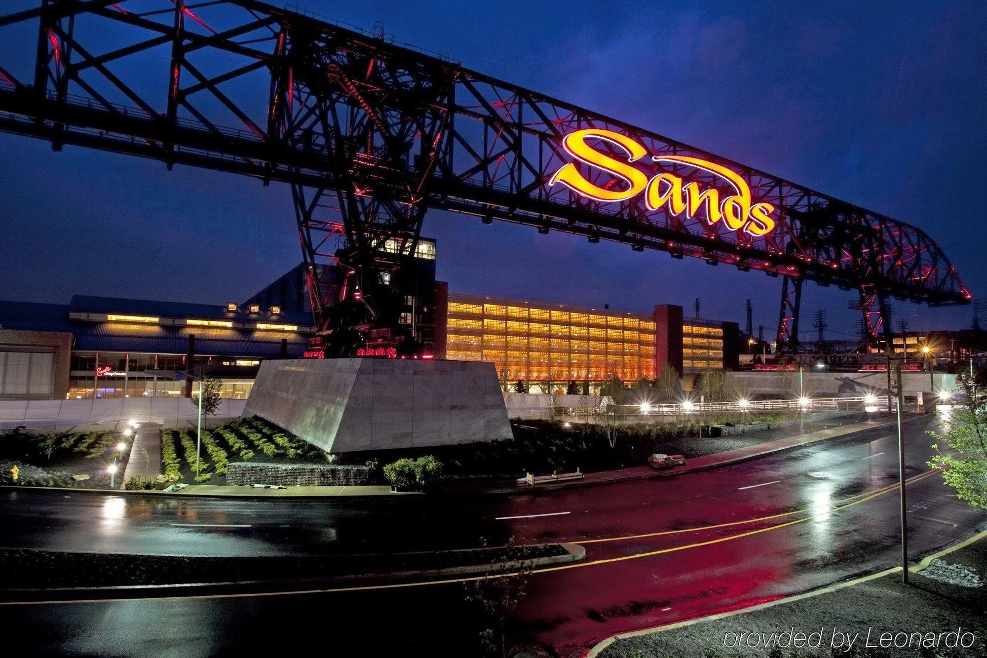 Night view of the Sands hotel and casino with a large illuminated sign reading 'Sands' on a metal bridge structure. The building is lit with yellow lights, and the scene shows a wet road in the foreground with streetlights and reflections on the pavement.