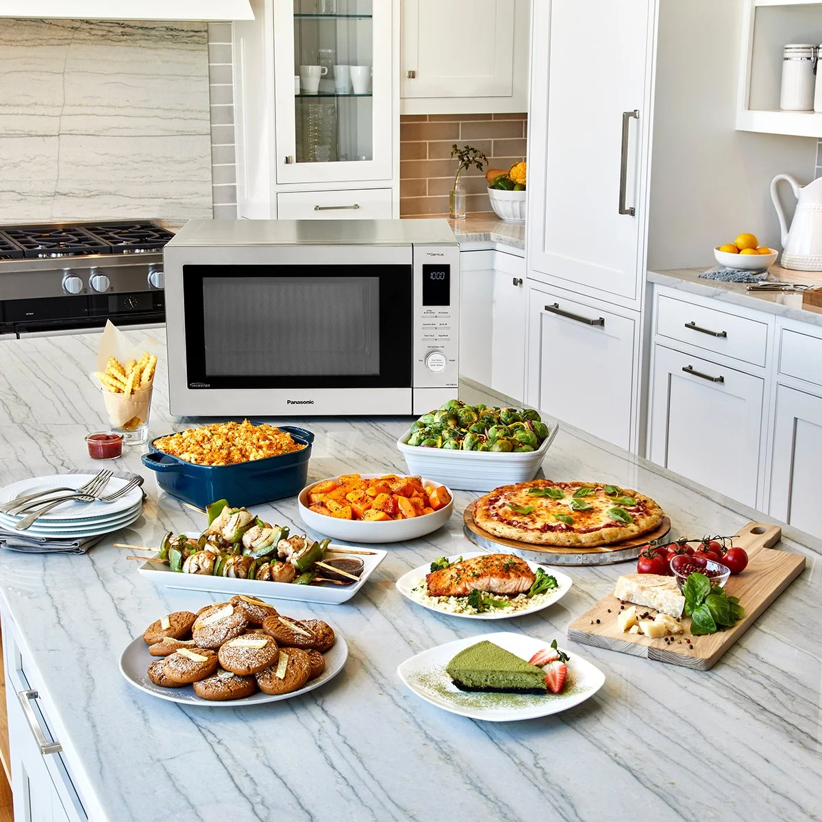 A kitchen countertop with various dishes including a pizza, a plate of cookies, a salmon fillet, a green cake slice, roasted vegetables, Brussels sprouts, and a bowl of tomatoes. There is a microwave oven and kitchen cabinets in the background.
