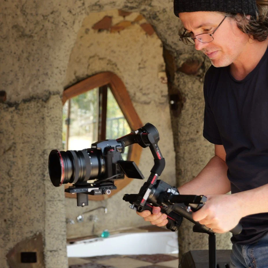 A man operating a camera stabilizer with a large professional camera attached, standing in a rustic room with a rounded window and a bathtub.