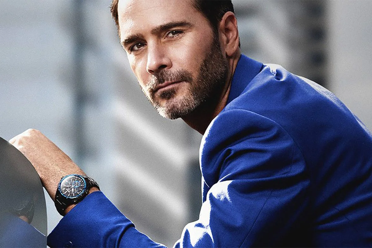 Man with a beard wearing a blue suit and a black watch, looking at the camera with a cityscape background.