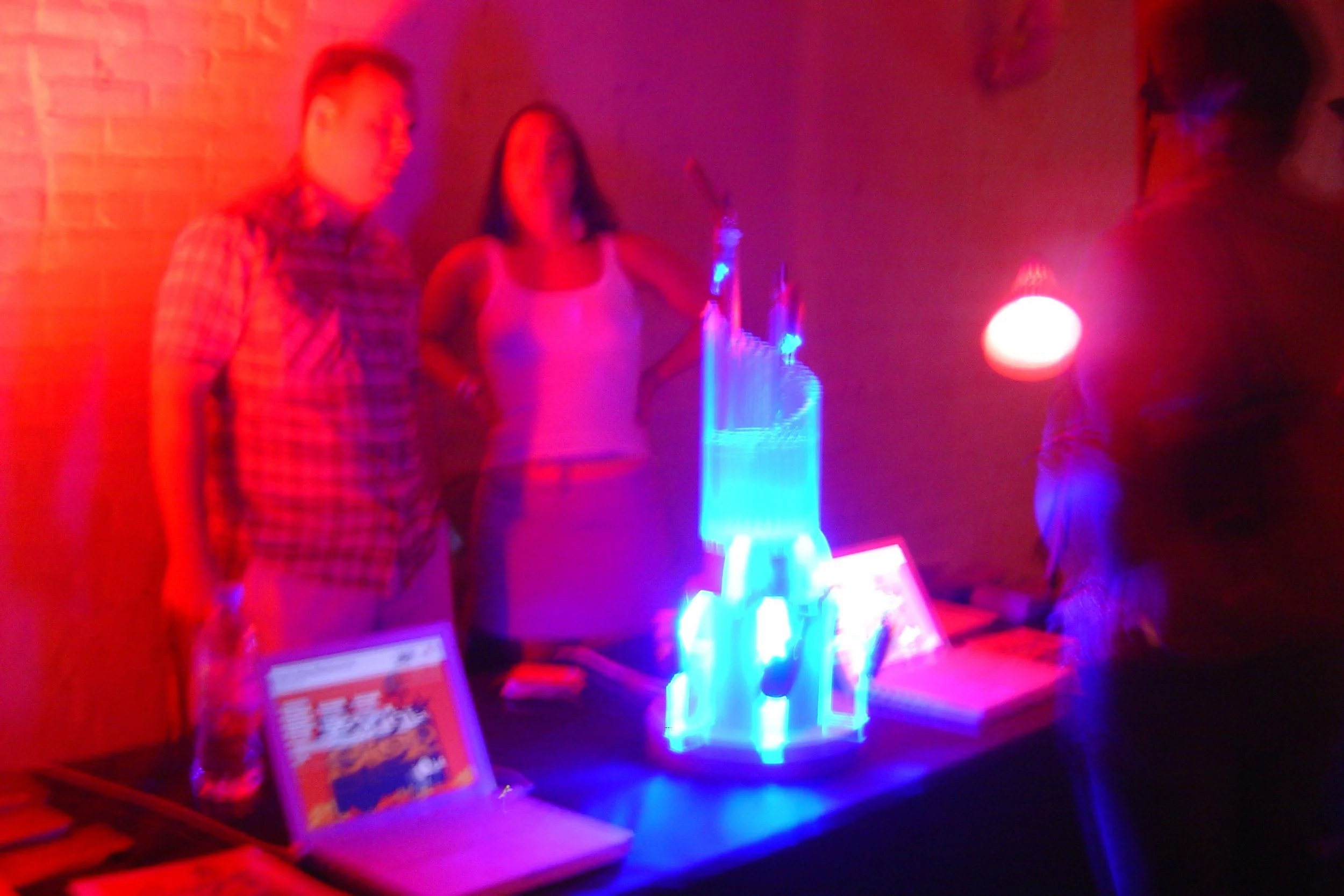 Three people standing around a table with illuminated objects and displays in a dimly lit room with red and blue lighting.