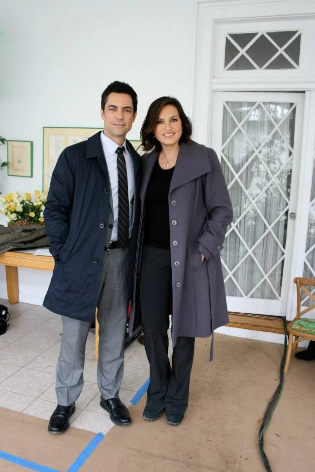 A man and a woman standing side by side indoors, smiling at the camera. The man is wearing a dark coat, light gray pants, and black shoes. The woman is wearing a dark coat, black pants, and black shoes. They are standing in front of a door with a win