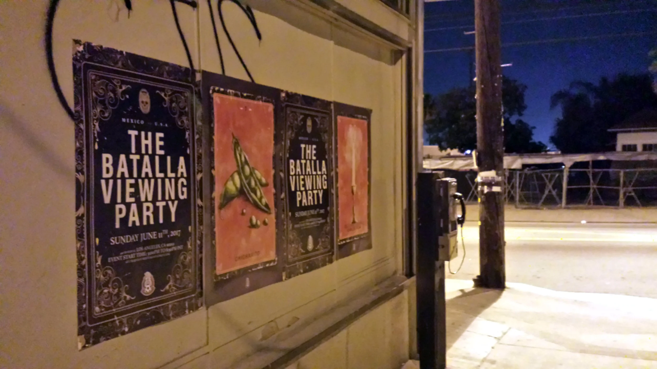 Three posters on a beige wall, with two promoting The Batalla Viewing Party and the middle one featuring a green butterfly illustration. Night scene with a street and telephone pole in the background.