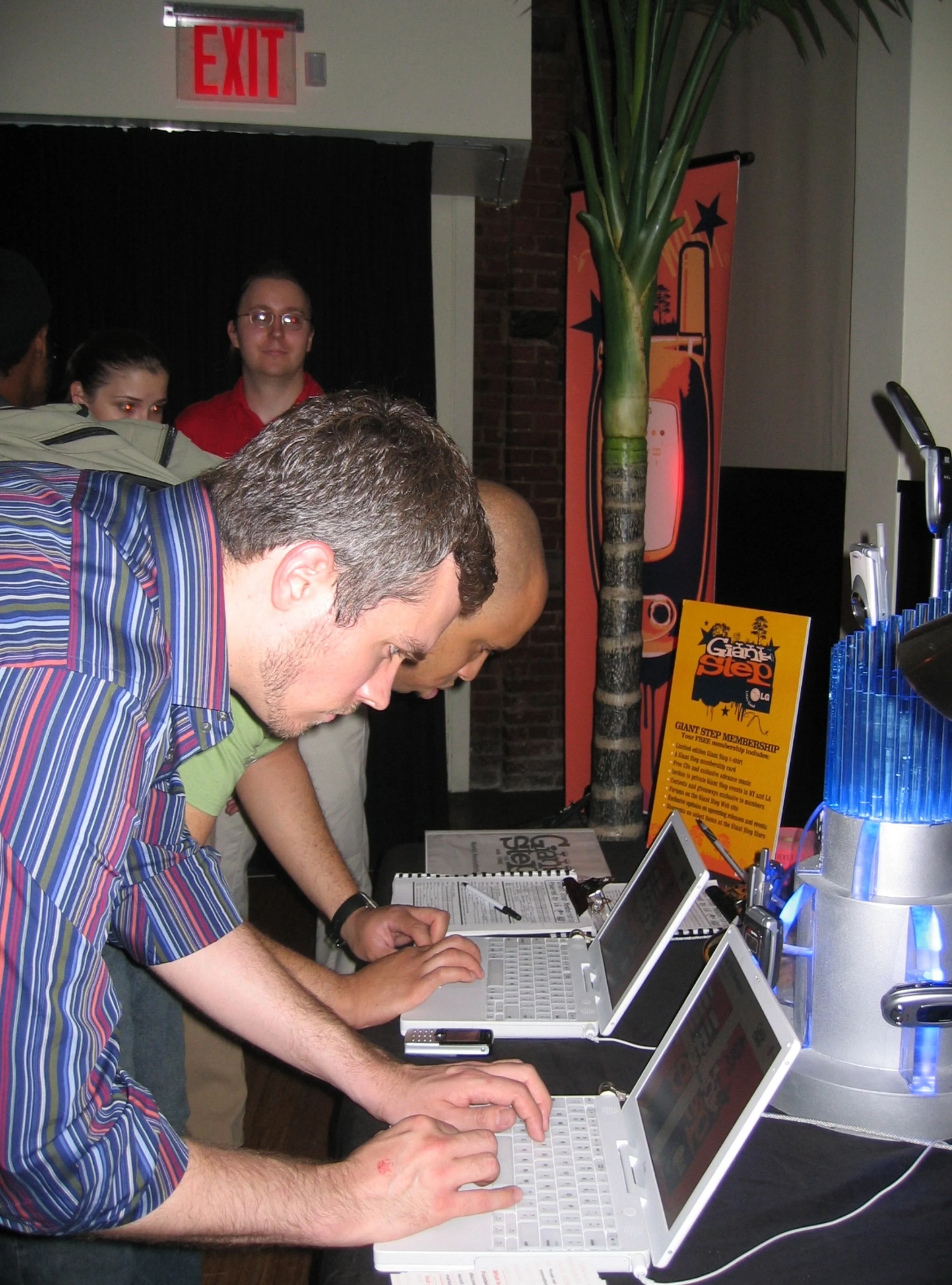 People standing at a table with laptops at a technology event, with promotional signs and a large palm tree in the background.