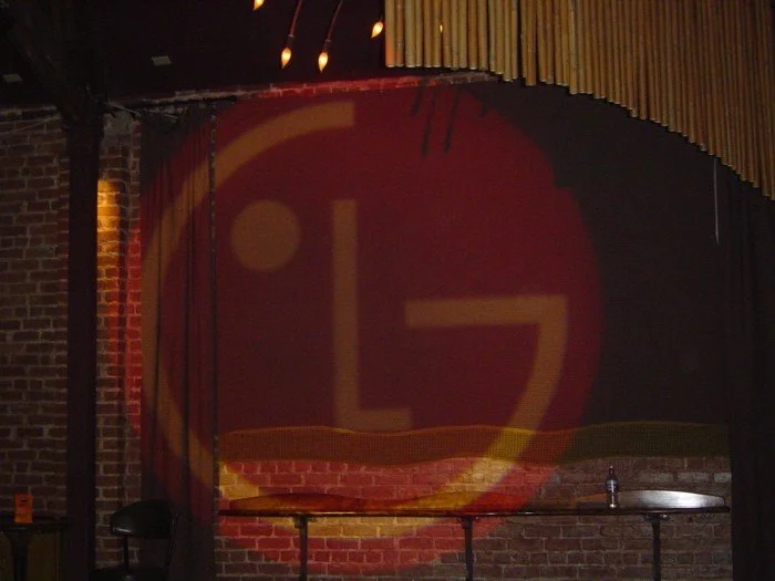 Stage with a brick backdrop, partially obscured red curtain, and a projected logo with the letters 'L7' inside a circle.