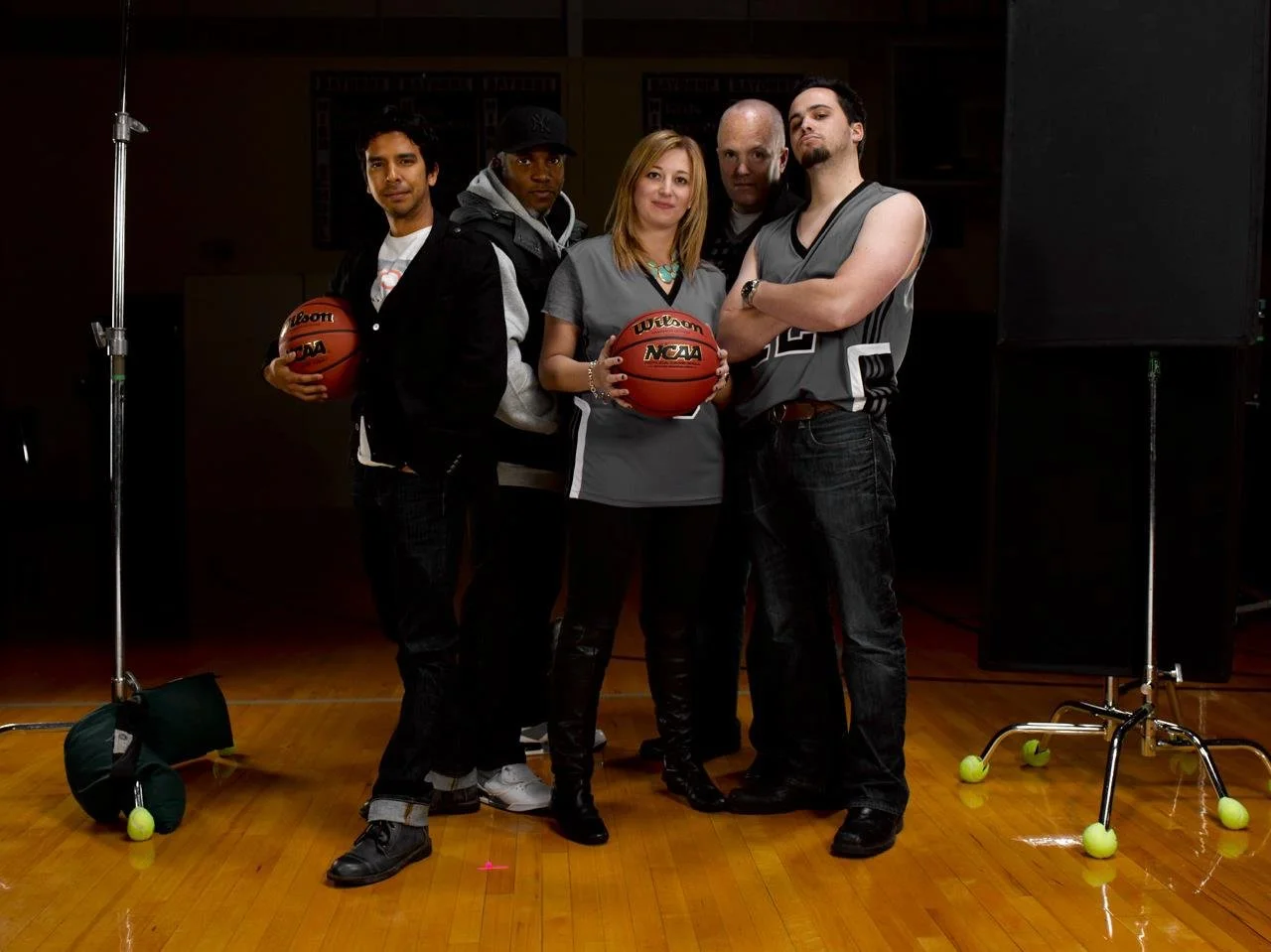 Group of five people standing on a gymnasium court, two holding basketballs, wearing casual and sports clothing, with tennis balls on the floor, and gym equipment visible.