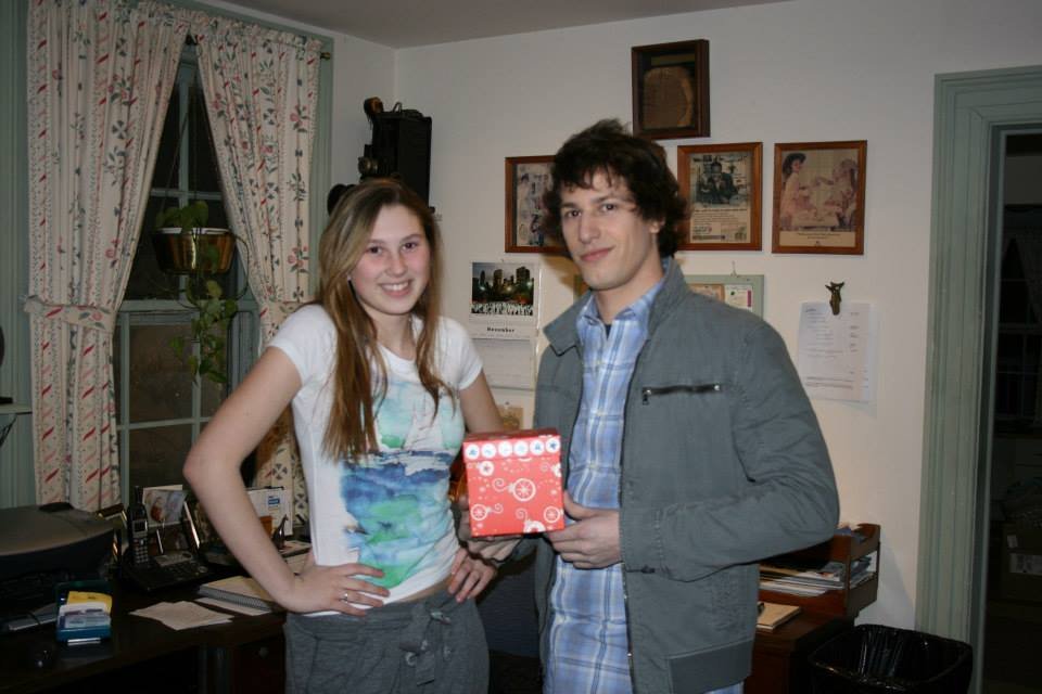 A young woman and a young man standing inside a room, smiling at the camera. The man is holding a small red gift box with white decorations. The room features a window with floral curtains, framed pictures on the wall, and an office desk with various