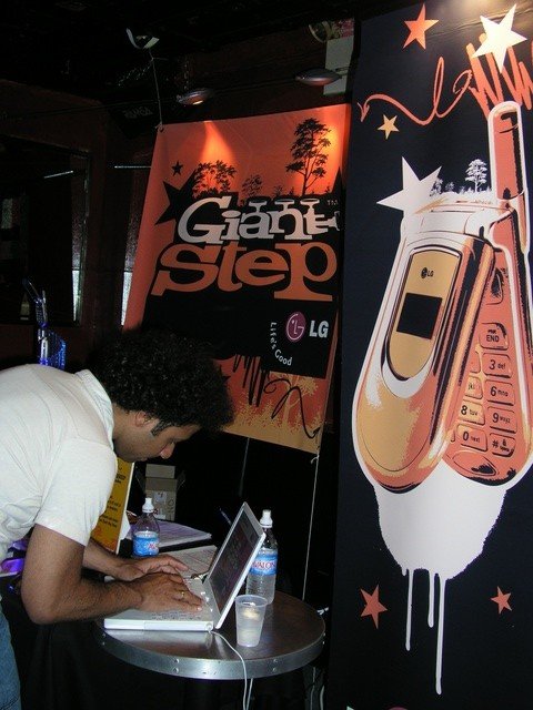 Young man using a laptop at a table with water bottles, in front of a promotional poster for LG featuring a flip phone and colorful stars.