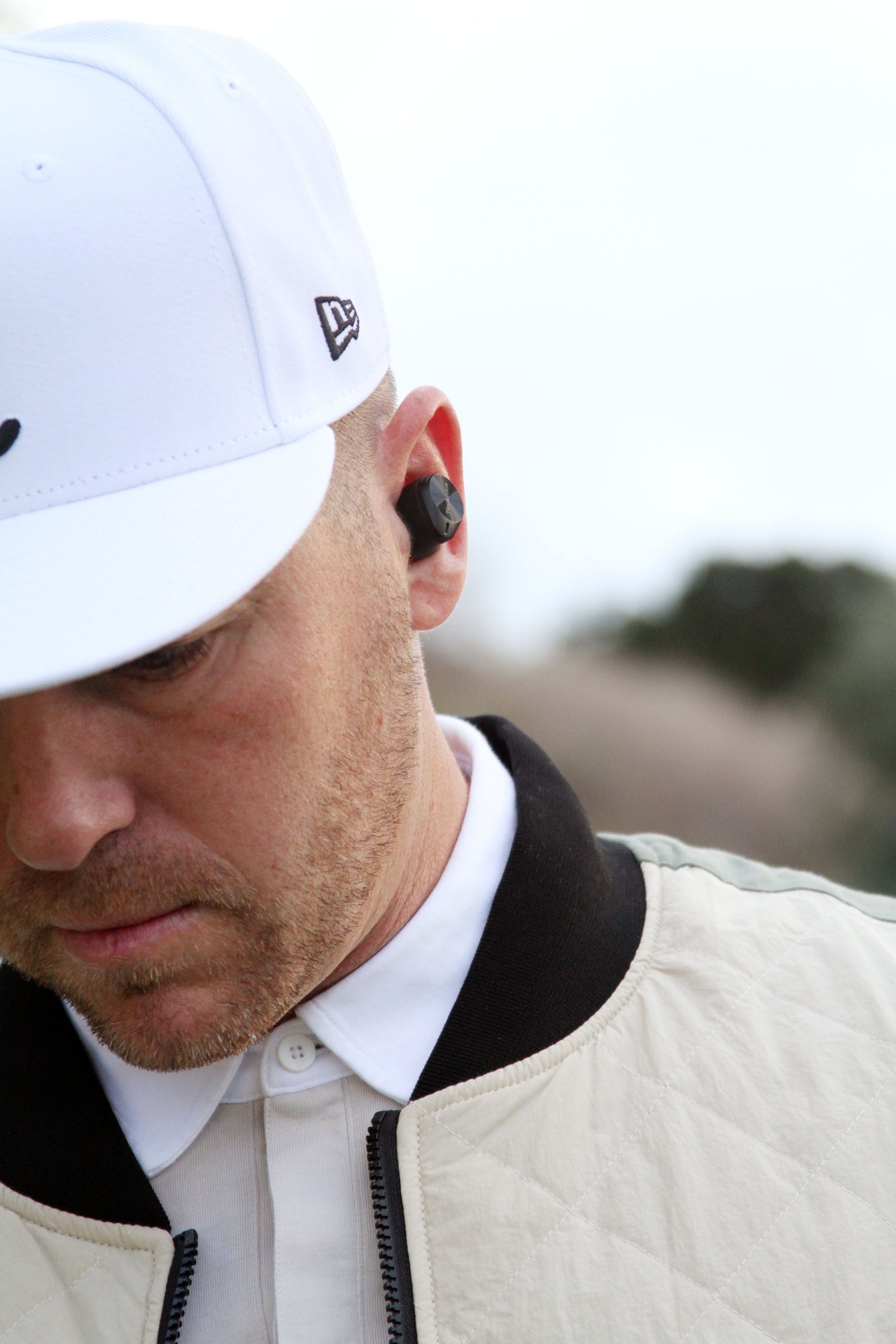 A man wearing a white cap, white jacket, and wireless in-ear headphones, looking downward with a focused expression.