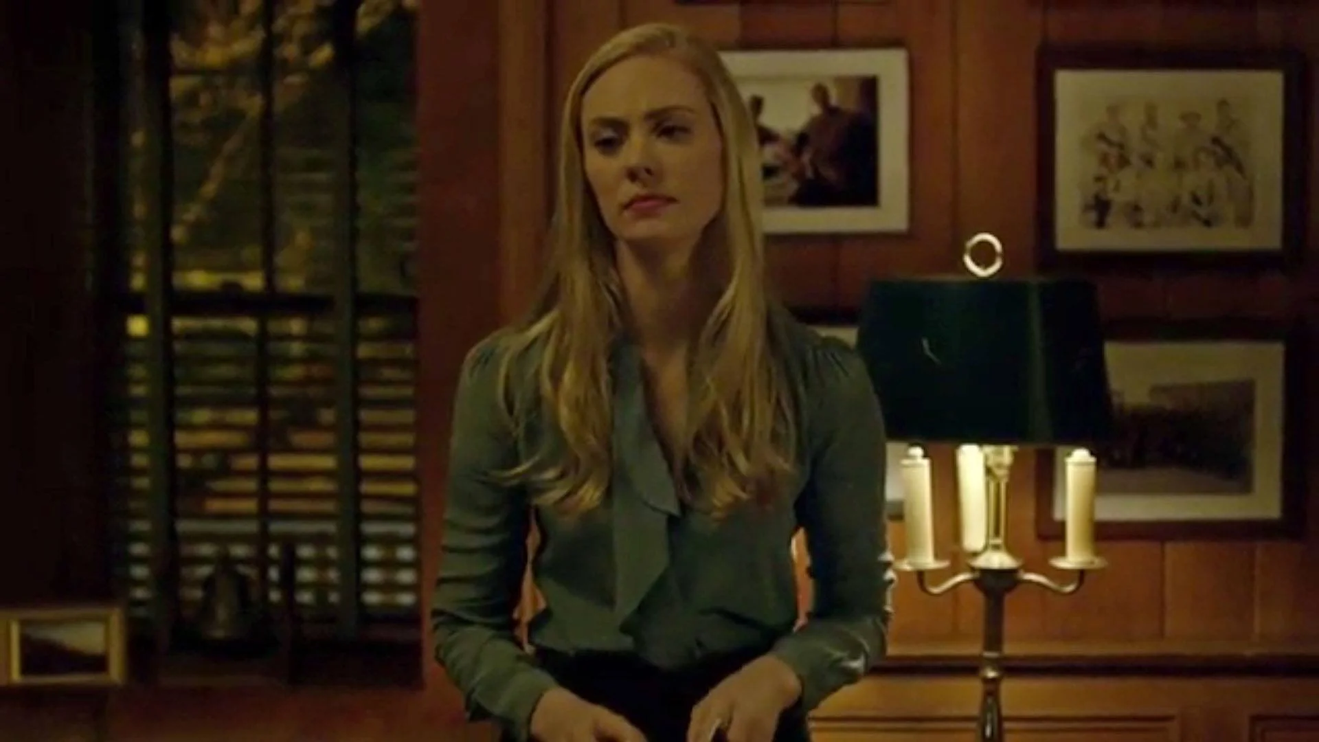 A woman with long blonde hair, wearing a gray blouse, stands in a dimly lit room with wooden walls. Behind her, there are framed pictures on the wall, a black lamp with a green shade, and a candelabra with three white candles.