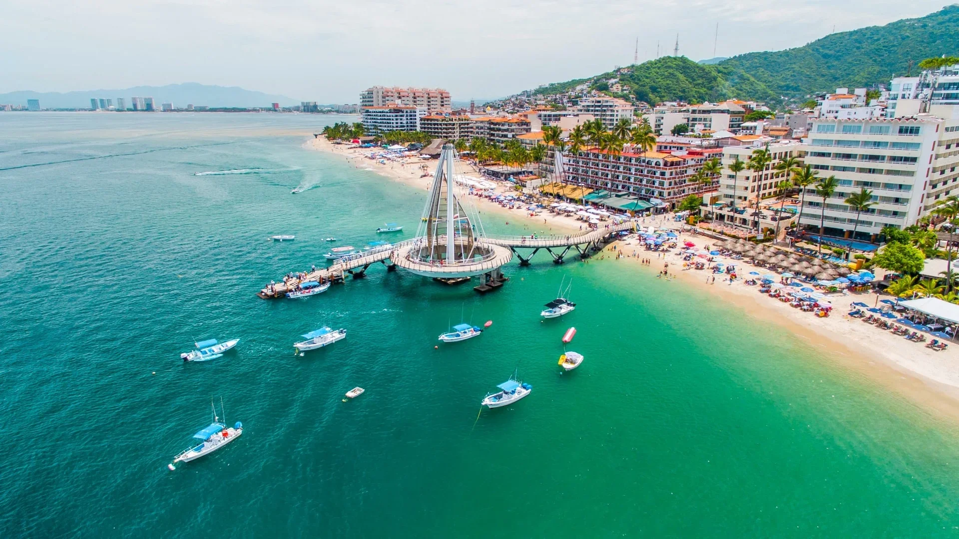 Aerial view of a beach with boats, a pier, high-rise hotels, palm trees, and a green hillside in the background.