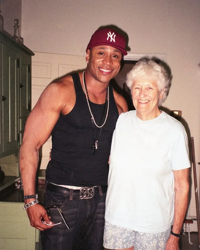 A young man wearing a red New York Yankees cap, a black sleeveless shirt, and jewelry standing beside an elderly woman with white hair in a white t-shirt and patterned shorts, both smiling indoors.