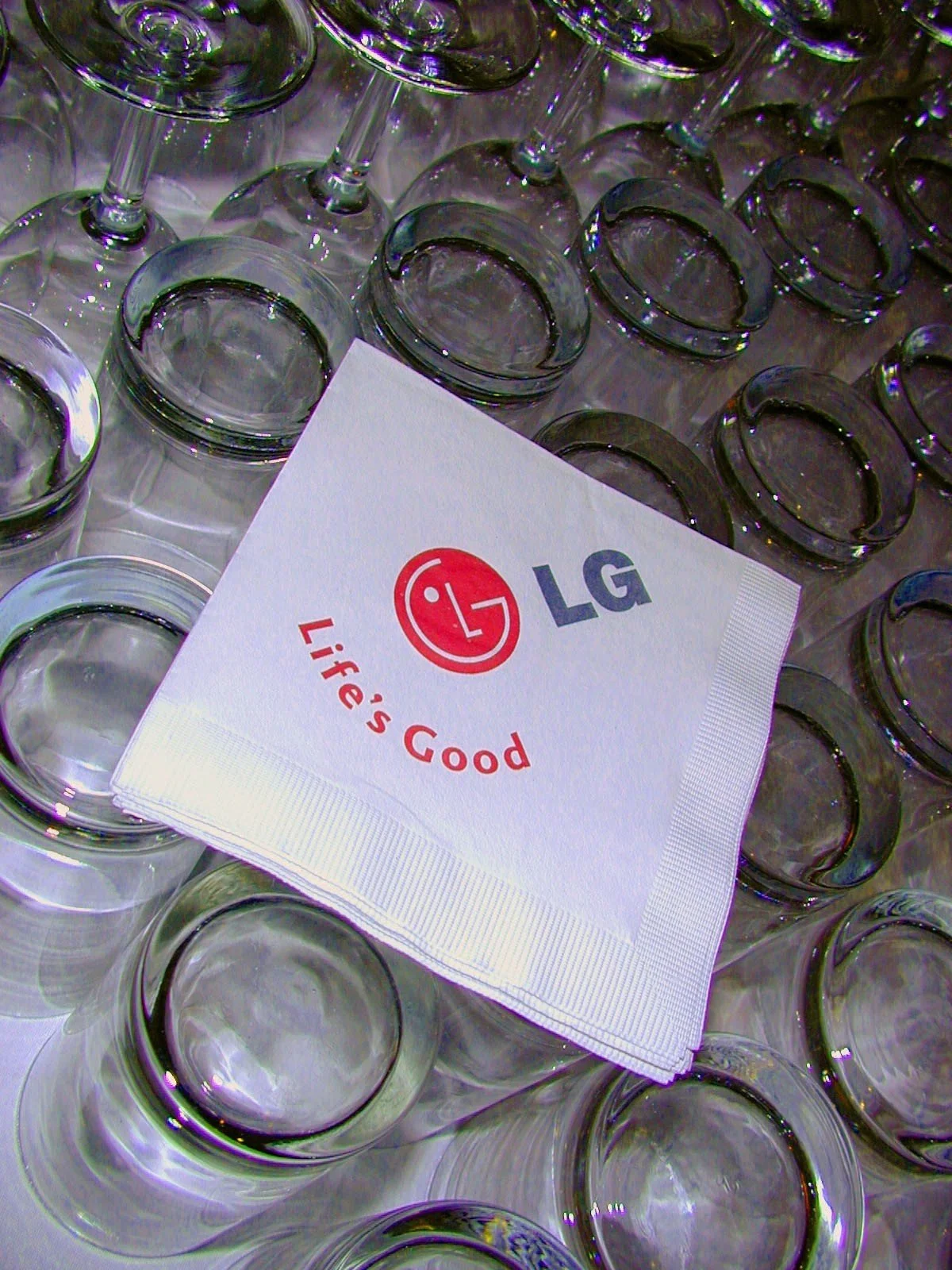 A stack of clear glass cups upside down, with a paper napkin on top featuring the LG logo, a smiley face, and the slogan 'Life's Good'.