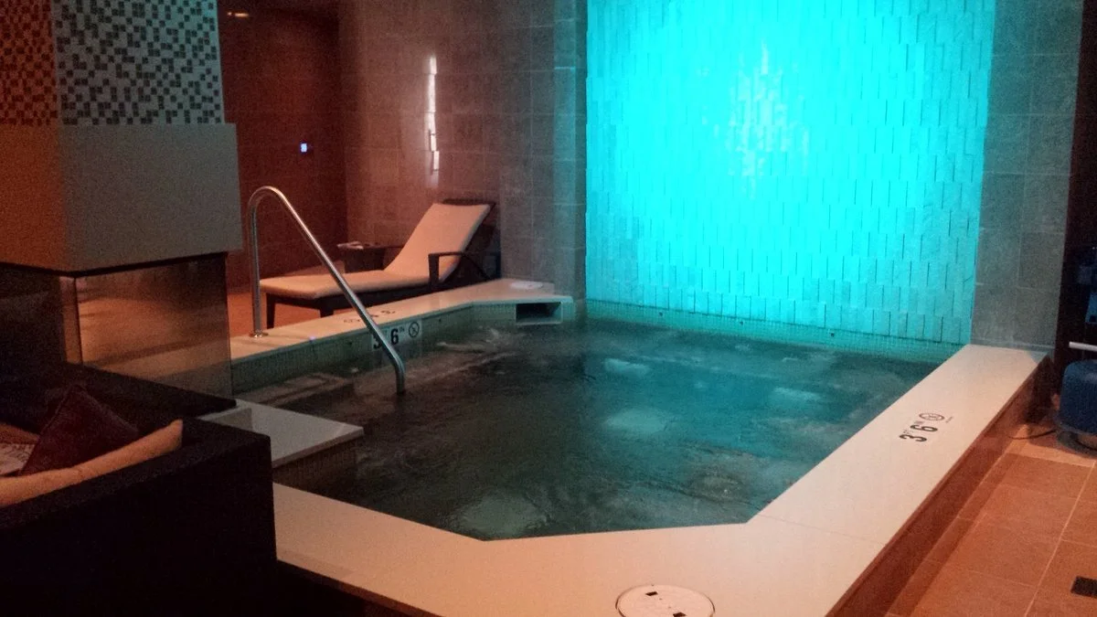 Indoor hotel spa with a small pool, illuminated blue waterfall feature, lounge chair, and tiled walls.
