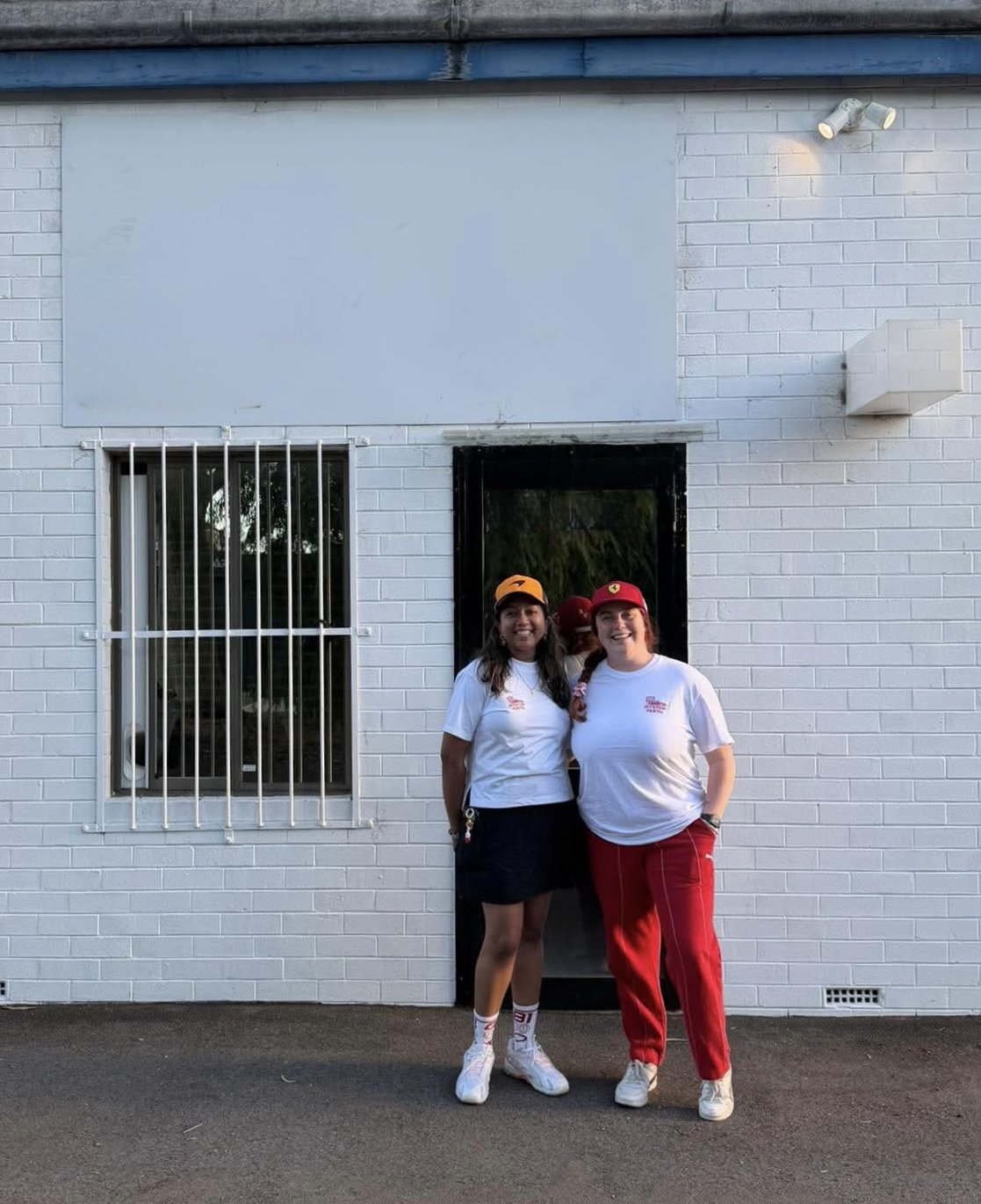 Two women standing in front of a white brick building, smiling. One woman is wearing a white t-shirt, black skirt, and an orange cap. The other woman is wearing a white t-shirt, red pants, and a red cap. Behind them is a black door and a window with security bars.
