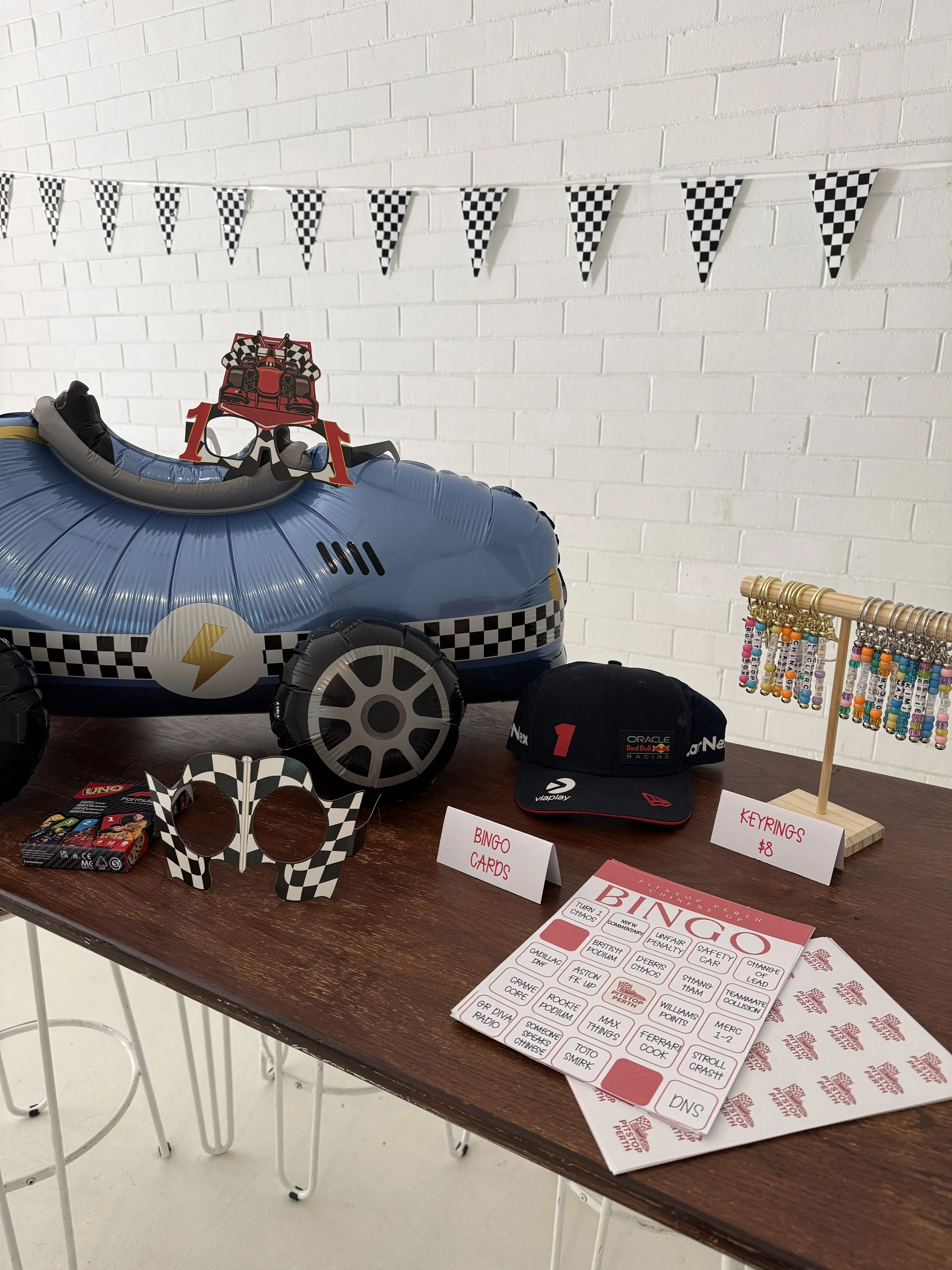 A wooden table displaying various racing-themed items and stationery. Items include a mascot beer hat, a bag with the Oracle Red Bull Racing logo, a toy race car, a checkered headband, and a bunch of beaded keychains hanging on a small display. There are also signs indicating prices for keyrings and bingo cards, a quiz sheet about the history of racing, and a racing-themed bingo game card. In the background, a white brick wall with a checkered flag banner and a small section of a black cap with a red number 1 logo.