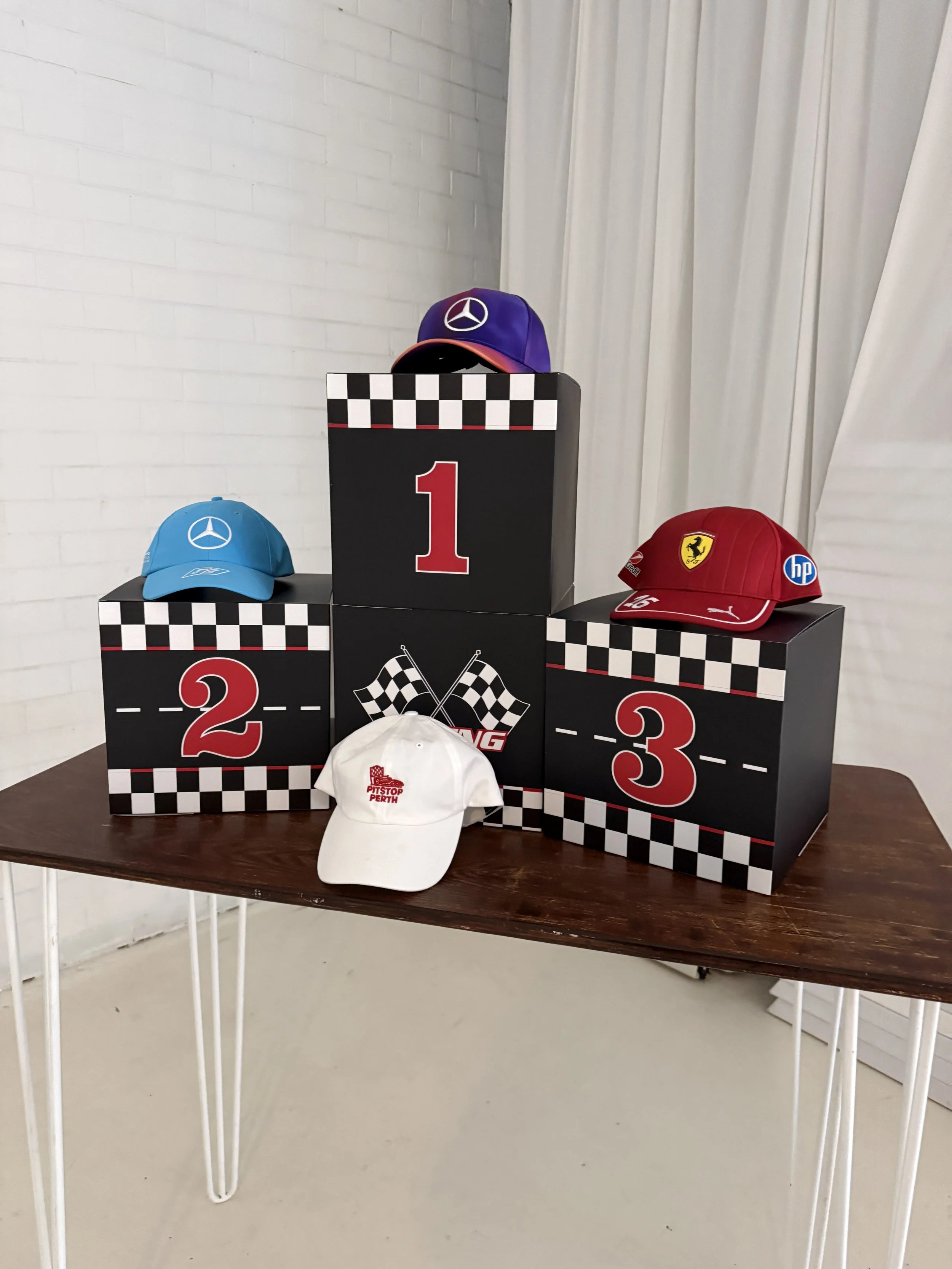 Race winners podium with three caps and a white hat on a brown table in front of a white brick wall and white curtains. The blue cap is on the second place podium, the purple cap on the first place, and the red cap on the third place podium. The white hat has a logo with red text.
