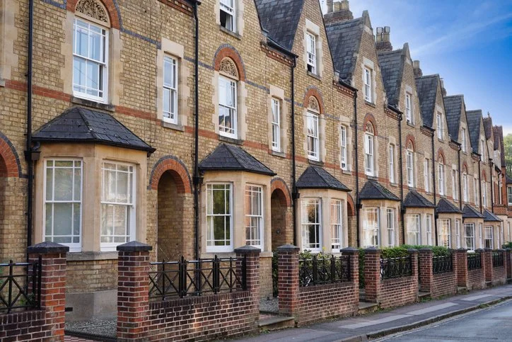 Row of attached brick townhouses with rounded bay windows, black fences, small front yards, and a sidewalk, under a blue sky.