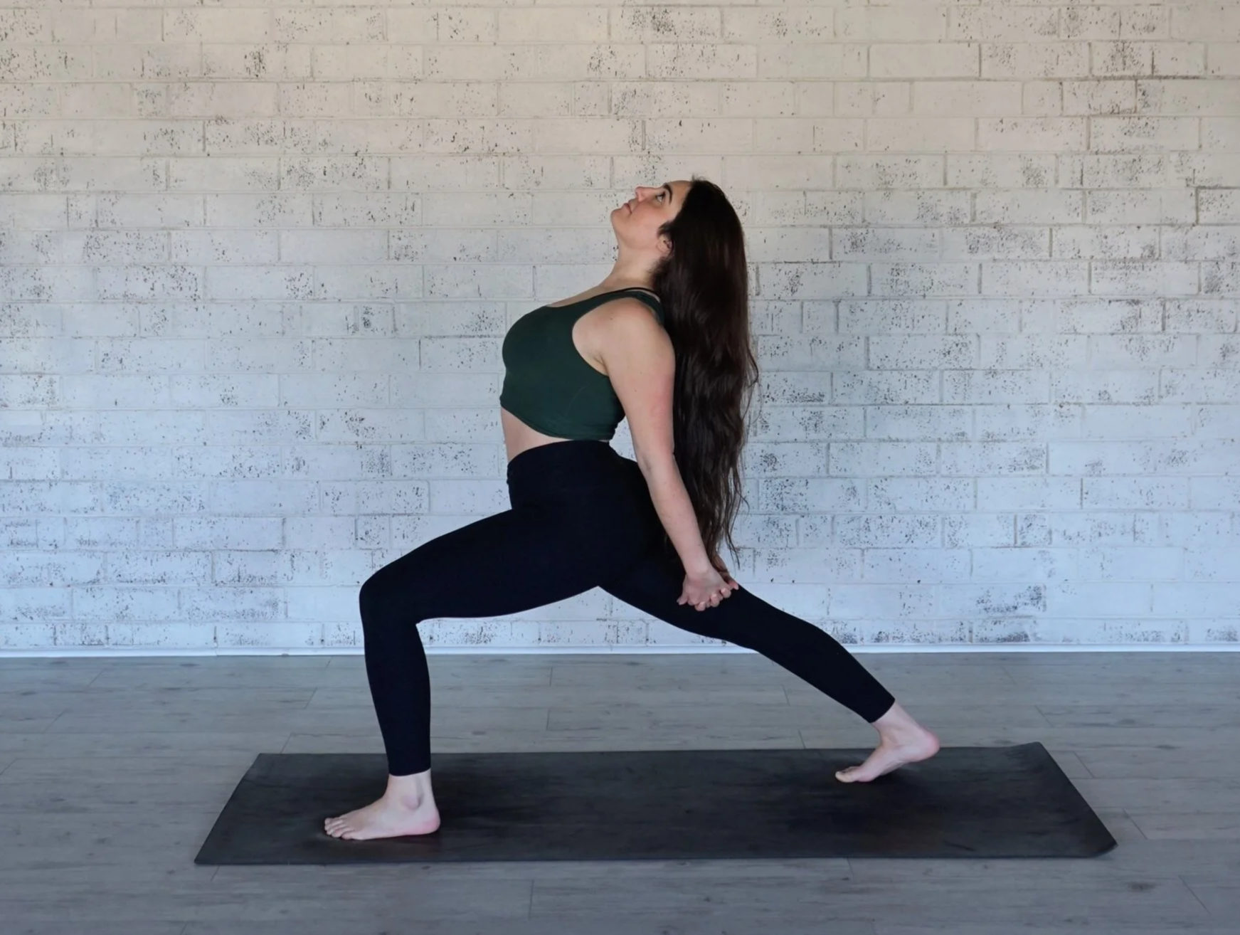 A woman practicing yoga in a lunge pose with arms extended upwards indoors near a white brick wall.