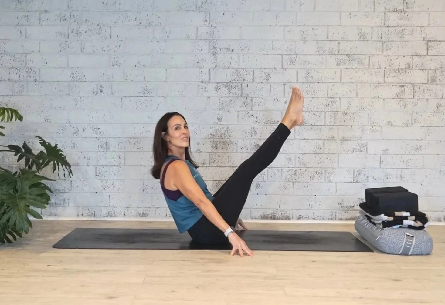 Woman in athletic clothing practicing yoga on a mat in a studio with a white brick wall, next to a plant and a pile of yoga props.
