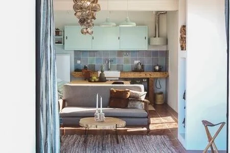 Cozy living room with a gray sofa, wooden coffee table, and a kitchen with pastel cabinets and a wooden countertop in the background.