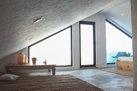 Bright attic bedroom with large triangular windows overlooking the ocean, modern furniture, and a sloped ceiling.