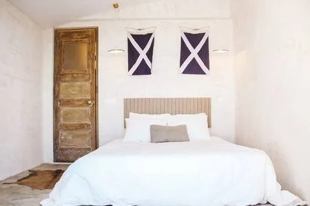 A bedroom with a white bed, two flags on the wall, a wooden door, and a small rug on the floor.