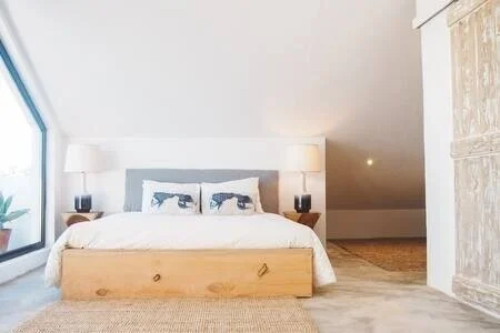 Bedroom with a large bed, blue and white pillows, two lamps on bedside tables, and a window with a view outside.
