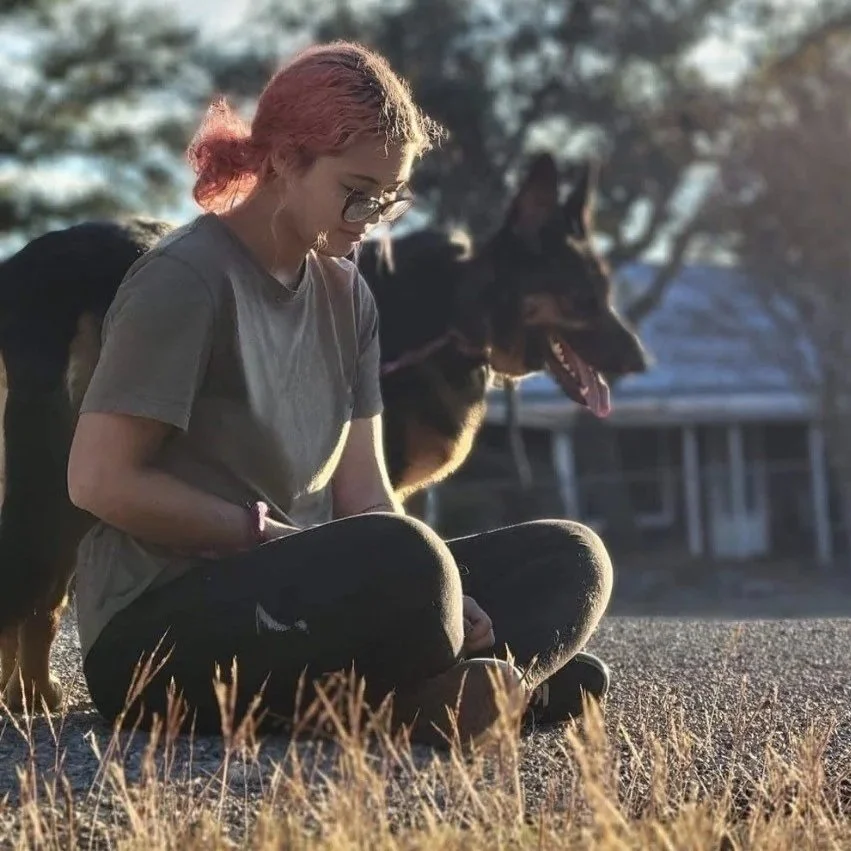 A young woman with pink hair, glasses, and a gray t-shirt sitting cross-legged outdoors at sunset, with a German Shepherd dog sitting behind her. Bonding with an aggressive dog.