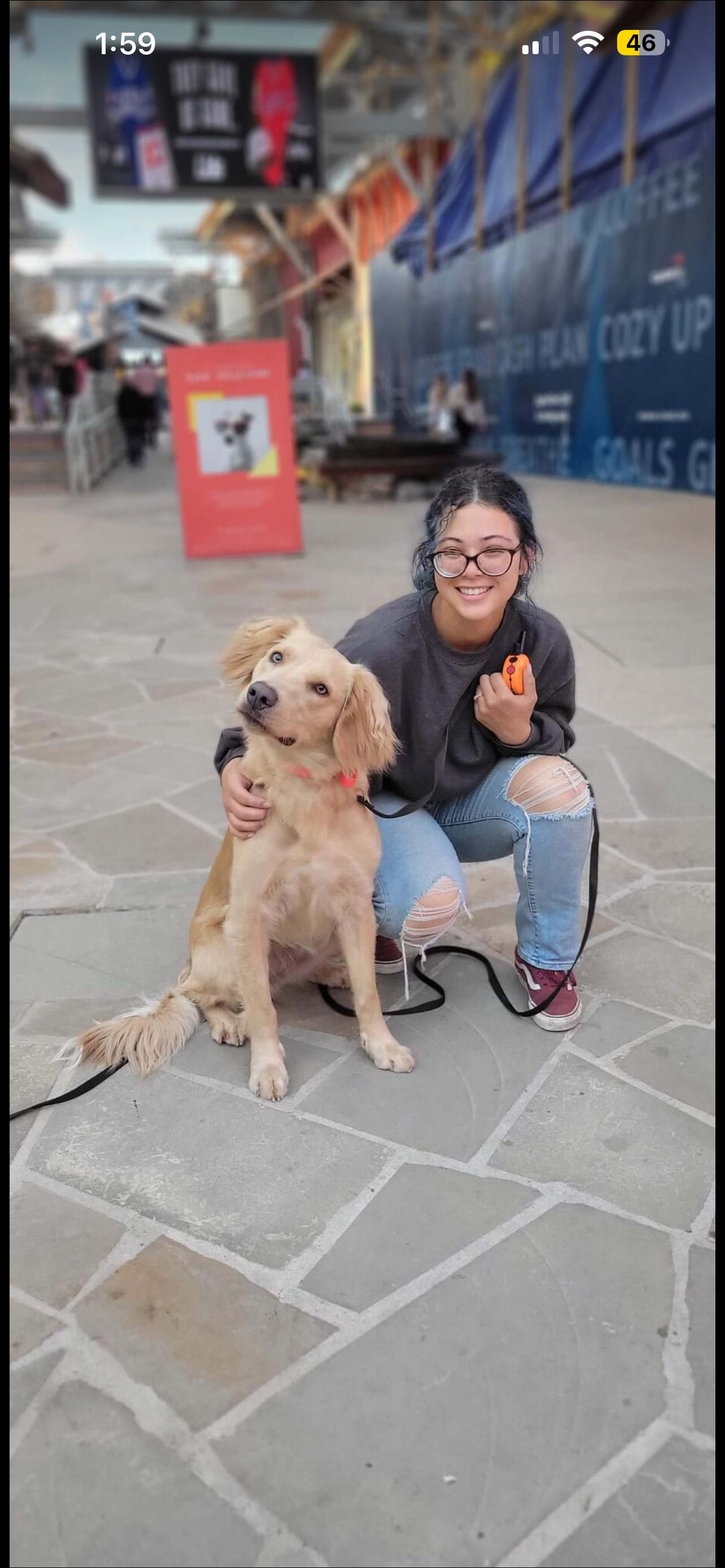 A young woman with glasses kneeling on a paved area, smiling, holding a small orange device in her right hand, with her left arm around a yellow retriever dog sitting beside her. The background shows an outdoor area with people, colorful structures, and a large sign