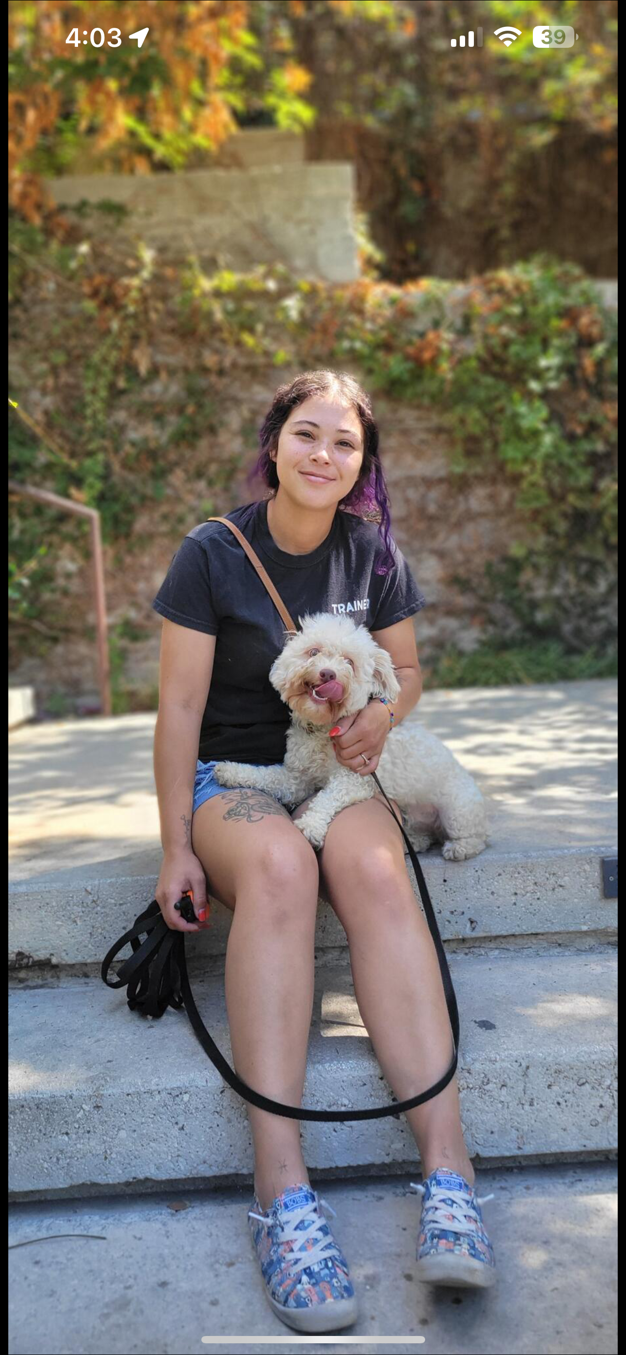 A woman with purple hair and tattoo on leg sitting on concrete steps outdoors, holding a smiling cream-colored dog with curly fur, in front of a rocky and leafy background.
