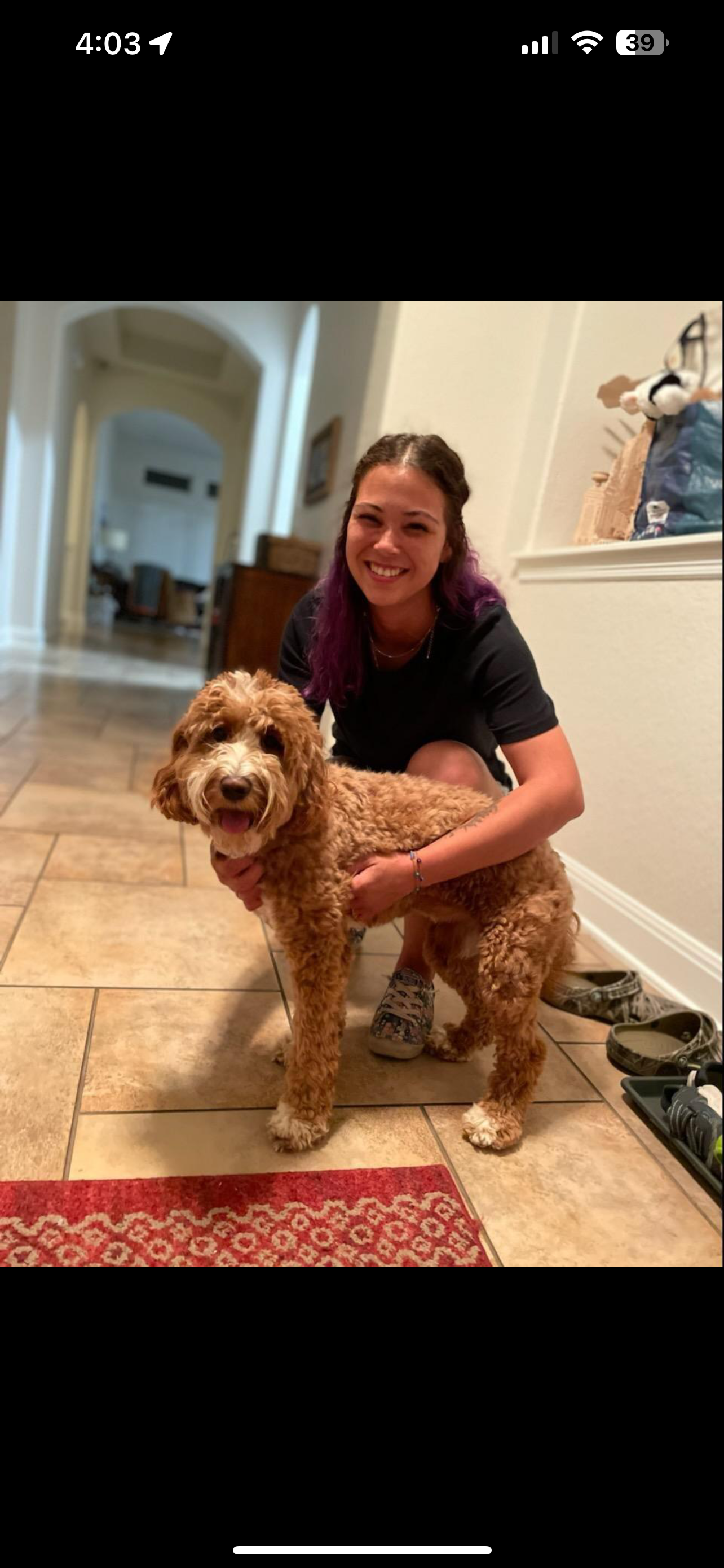 A woman with purple hair crouches down to hug a curly brown dog in a home with tiled floors and a hallway in the background. Saying goodbye to a dog who has just graduating from our dog training in San Antonio, Tx.