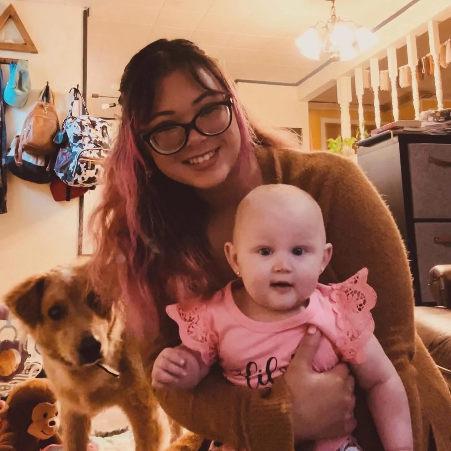 A woman with pink-tinted hair, glasses, and a brown sweater smiling while holding a baby girl with a pink outfit. There is a small dog with a brown and white coat standing beside them in a cozy indoor setting.