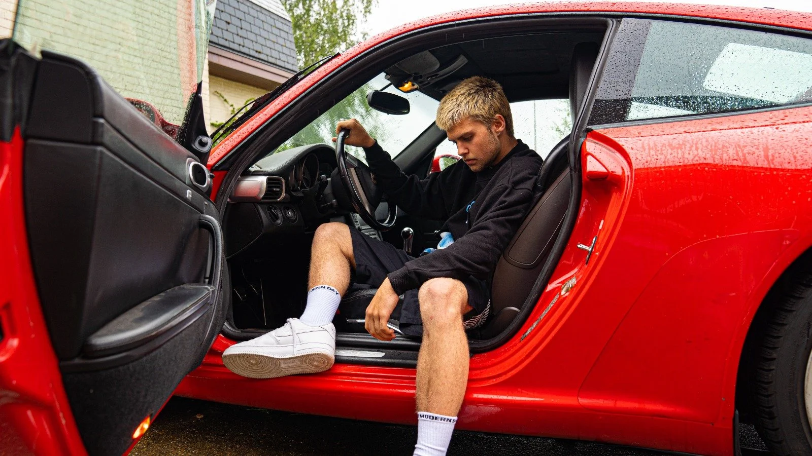 Man in zwart jack en korte broek zit in een rode sportauto, met de deur open, op een regenachtige dag, de man zit op de rand van de bestuurdersstoel met een hand op het stuur.