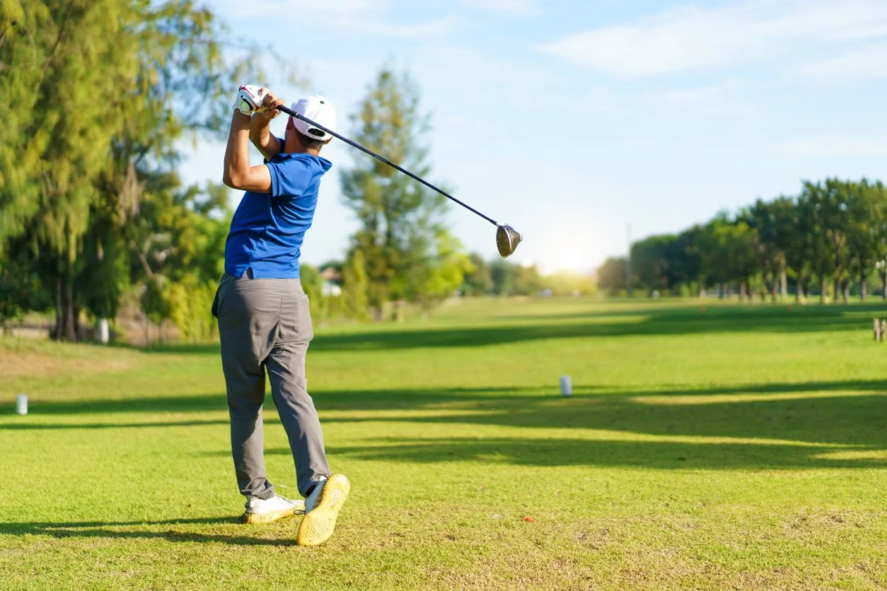 A man in a blue shirt and white cap playing golf on a sunny golf course.