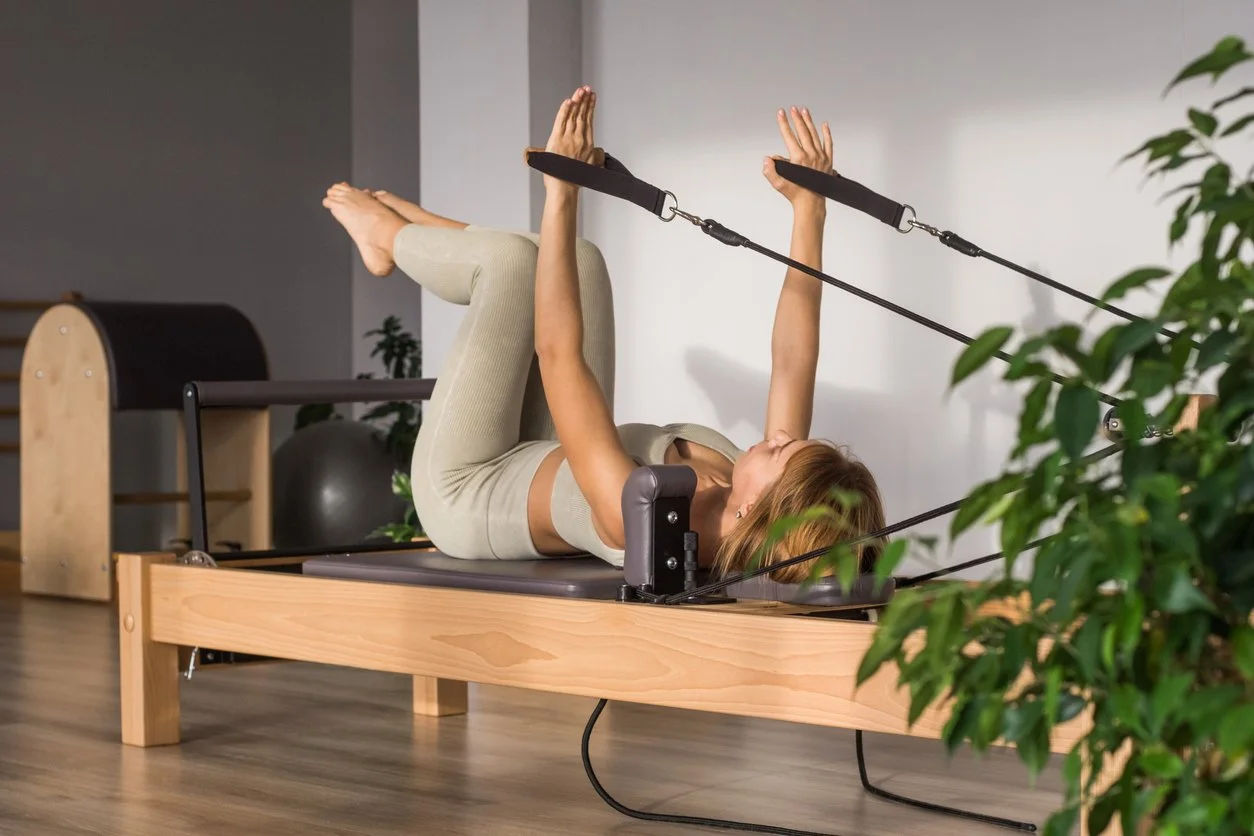 Woman exercising on a Pilates reformer with resistance straps, lying on her back with legs bent and arms raised.