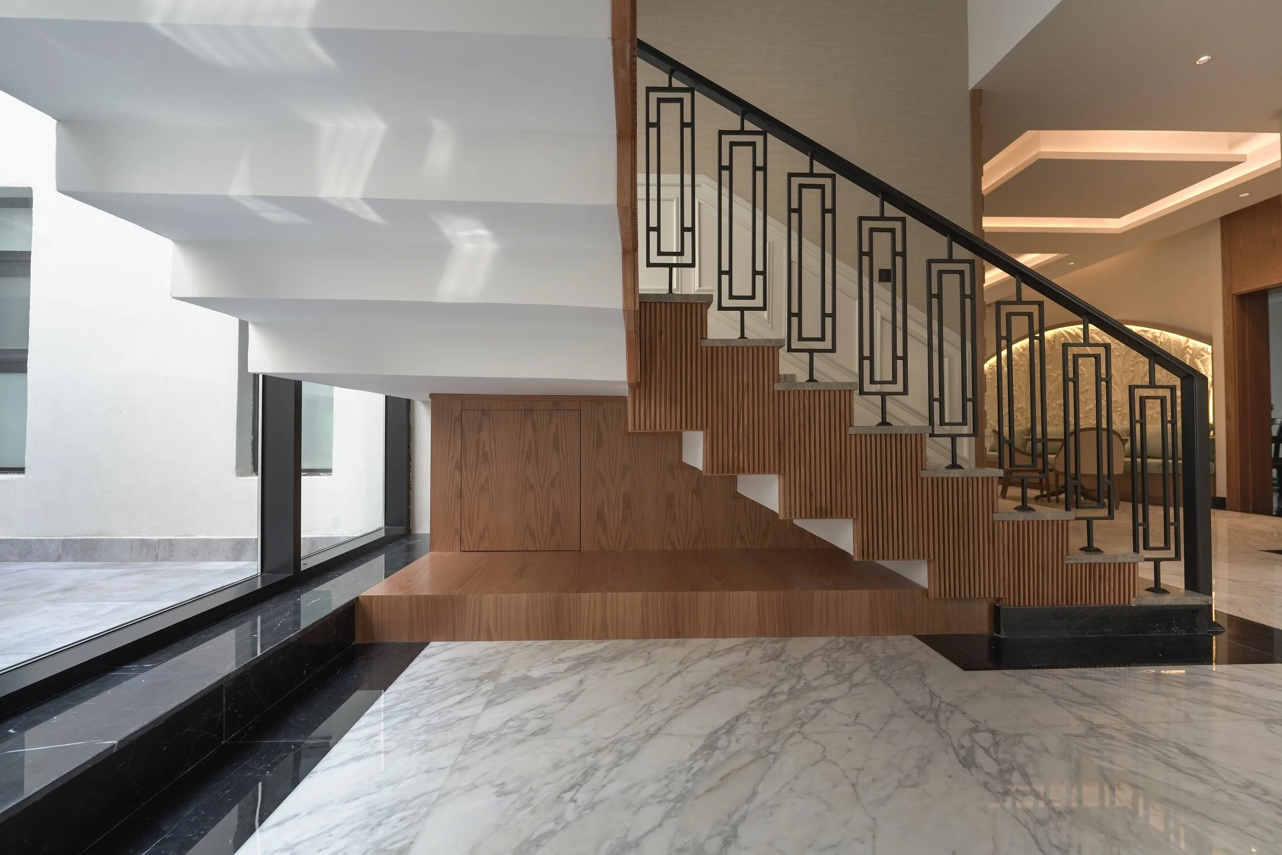 Interior of a modern building featuring a staircase with wooden steps and a decorative metal railing, polished marble flooring, large windows, and ambient ceiling lighting.