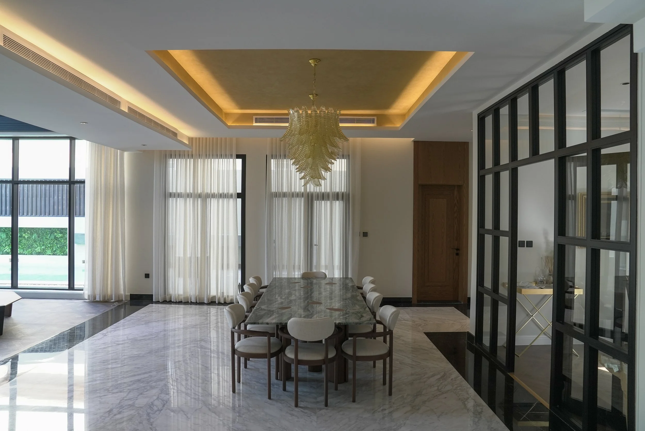 Elegant dining area with a marble table, surrounded by eight white chairs, modern chandelier, large windows with sheer curtains, and decorative black-framed glass panel wall.